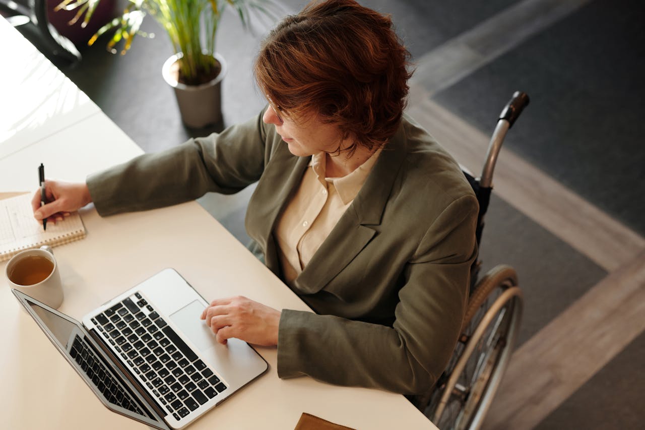 وظفني - لذوي الإعاقة Top view of a businesswoman in a wheelchair working at her desk with a laptop and notebook.